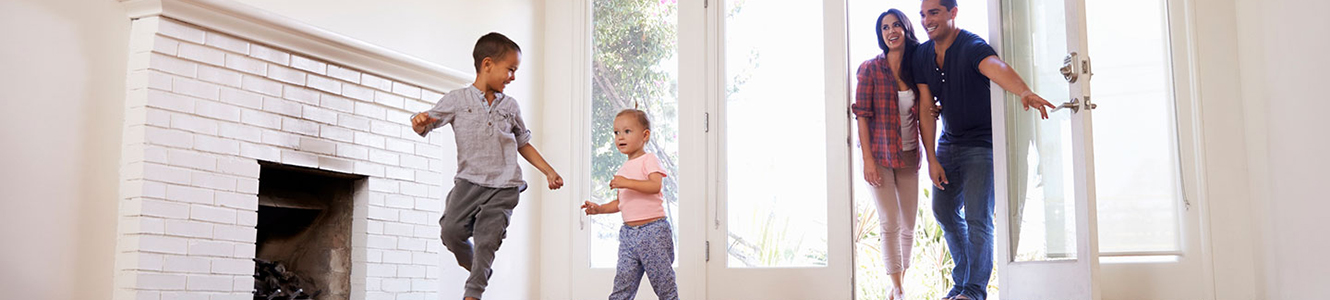 Mom and dad walking in front door of newly purchased home. Young son and daughter ahead of them running and smiling in living room. 